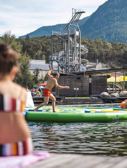 Personen spielen auf einem aufblasbaren Hindernisparcours in einem See, während im Vordergrund zwei Frauen auf dem Steg sitzen, umgeben von Bäumen und Bergen.