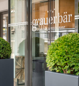 Entrance to "Grauer Bär" restaurant and hotel, featuring large, round, green shrubs in gray planters next to a glass door that reflects the surroundings. Signs and reviews are posted on the window.