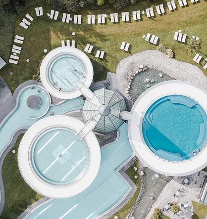 Luftaufnahme einer modernen Freibadanlage mit runden Becken, Wegen und zahlreichen Liegestühlen, die ordentlich auf dem grünen Rasen angeordnet sind.