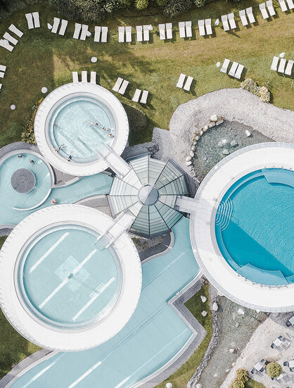 Luftaufnahme einer modernen Freibadanlage mit runden Becken, Wegen und zahlreichen Liegestühlen, die ordentlich auf dem grünen Rasen angeordnet sind.