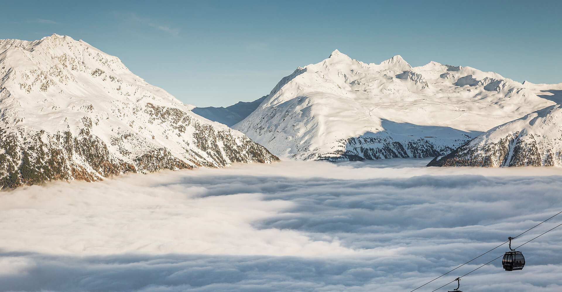 Snow-covered mountains rise from a thick blanket of clouds, while below to the right, under a bright blue sky, two mountain gondola cabins move along a cable.