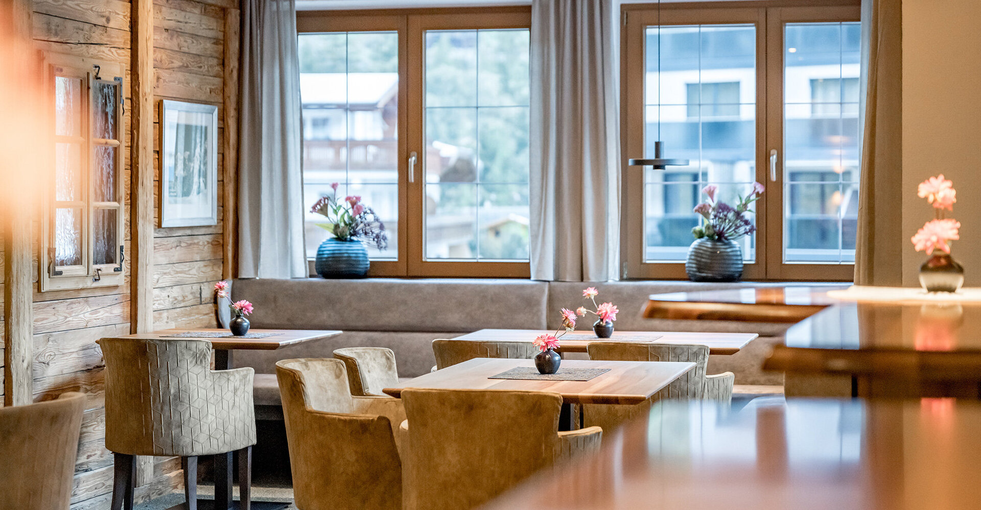 A cozy restaurant with wooden walls, large windows, beige chairs and tables decorated with small vases of flowers. Soft daylight floods the room, creating a warm and inviting atmosphere.
