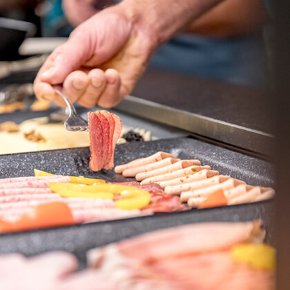 A person holding tongs selects a slice of cold cut from a platter featuring various types of cold cuts at a breakfast buffet.