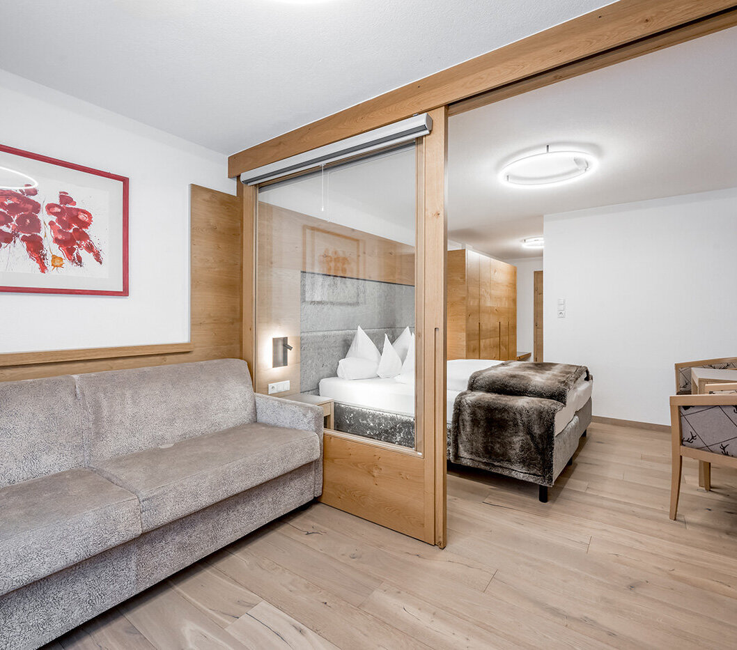 Modern hotel room featuring a gray sofa, a glass room divider with a wooden frame, a double bed with white bedding, a wall-mounted TV, a desk, wood accents and a picture on the wall.