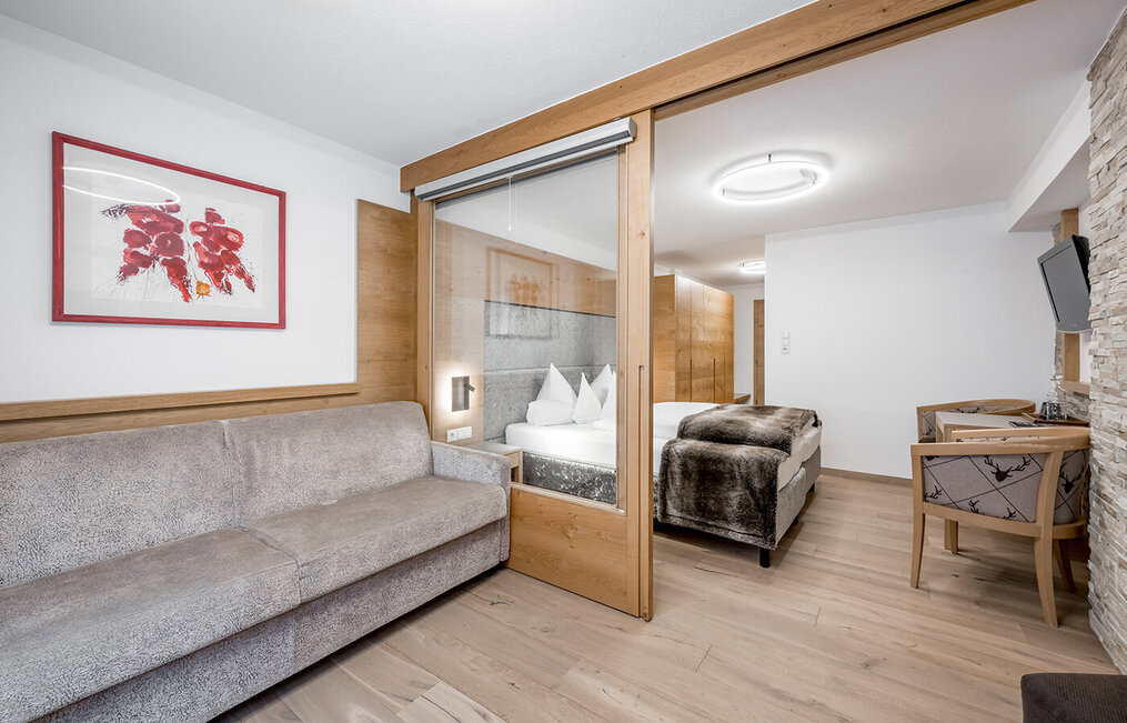 Modernes Hotelzimmer mit einem grauen Sofa, einem Raumtrenner aus Glas mit Holzrahmen, einem Doppelbett mit weißer Bettwäsche, einem an der Wand montierten Fernseher, einem Schreibtisch, Holzakzenten und einem Bild an der Wand.