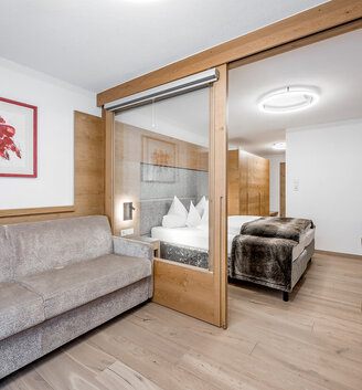 Modern hotel room featuring a gray sofa, a glass room divider with a wooden frame, a double bed with white bedding, a wall-mounted TV, a desk, wood accents and a picture on the wall.