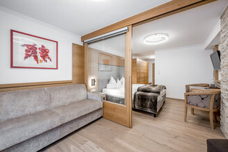 Modern hotel room featuring a gray sofa, a glass room divider with a wooden frame, a double bed with white bedding, a wall-mounted TV, a desk, wood accents and a picture on the wall.