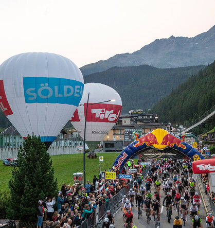 A huge number of cyclists start the race under a Red Bull arch, watched by a large crowd and flanked by hot-air balloons and mountains in the background.
