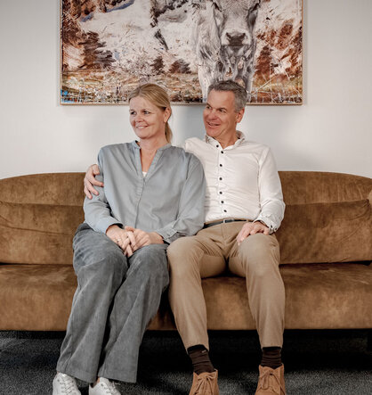 A smiling man and a smiling woman are sitting close together on a brown sofa in a modern room; a painting featuring a cow hangs on the wall behind them.