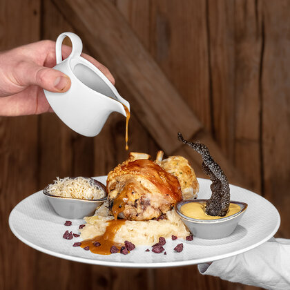 A hand pours sauce onto a plate, set against a rustic wooden background, containing a pork knuckle, rice, sauce in a small bowl and side dishes.