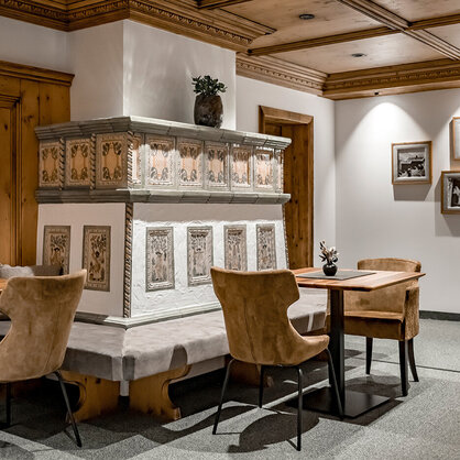 Cozy lounge with wooden tables and light-brown chairs, featuring a decorative ceramic stove, wood-paneled walls and framed photos on a white wall. Warm lighting creates an inviting atmosphere.