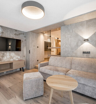 Modern living room featuring a beige sofa, a round wooden coffee table, a wall-mounted TV, pendant lights and wooden flooring. The textured walls are adorned with a white floral pattern. In the background, there is an unobstructed view of the sleeping area.