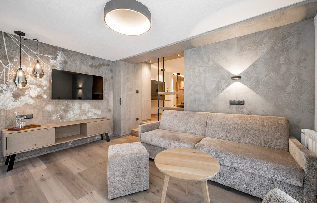 Modern living room featuring a beige sofa, a round wooden coffee table, a wall-mounted TV, pendant lights and wooden flooring. The textured walls are adorned with a white floral pattern. In the background, there is an unobstructed view of the sleeping area.