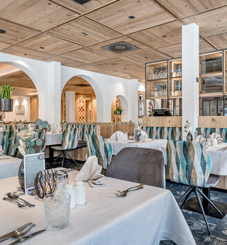 A modern restaurant featuring a wooden ceiling, white tablecloths, neatly set tables, patterned booth seating, glass partitions and soft lighting, creating an inviting and elegant dining atmosphere.
