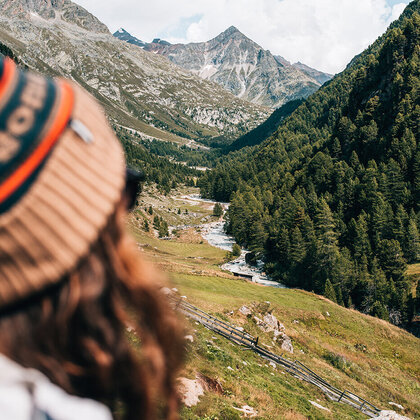 A person wearing a brown hat looks out at a picturesque mountain landscape featuring a river, lush green trees and rocky peaks under a partly cloudy sky.