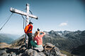 Zwei Wanderer ruhen sich neben einem großen Holzkreuz auf einem Berggipfel aus und genießen den malerischen Ausblick auf die schroffen, weit entfernten Gipfel unter einem strahlend blauen Himmel.