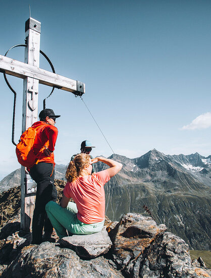 Two hikers are resting beside a large wooden cross on a mountain peak, enjoying the picturesque view of rugged, distant summits under a brilliant blue sky.
