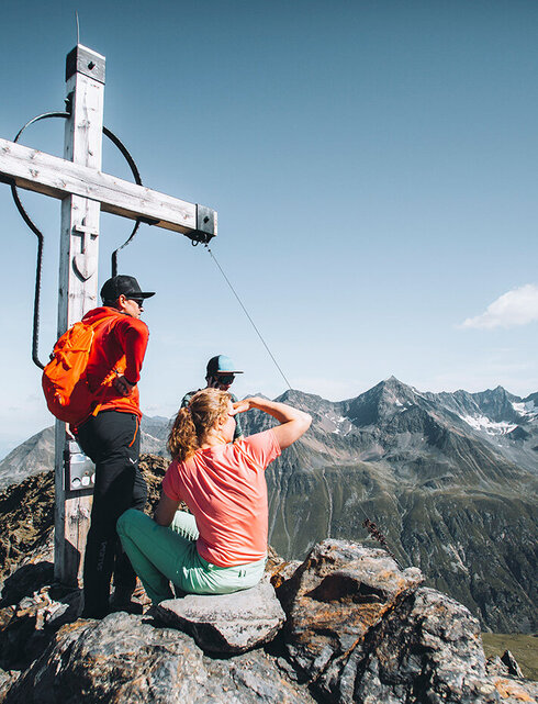 Two hikers are resting beside a large wooden cross on a mountain peak, enjoying the picturesque view of rugged, distant summits under a brilliant blue sky.