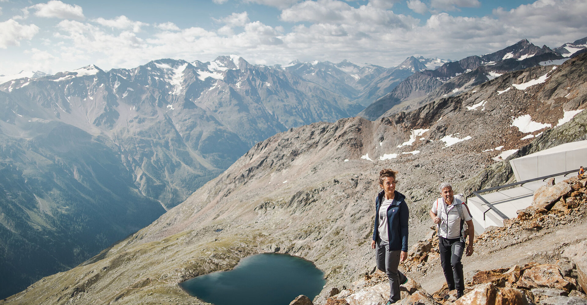 Zwei Wanderer gehen auf einem felsigen Bergpfad, im Hintergrund sind ein herzförmiger Bergsee und Gipfel zu sehen, während der Himmel teilweise bewölkt ist.