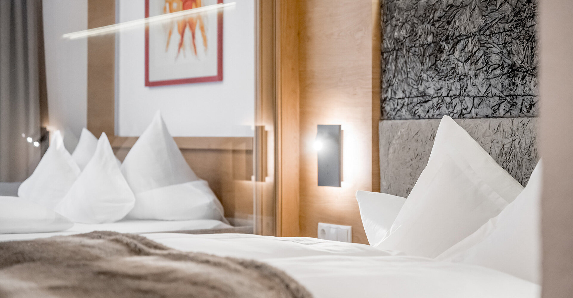 Ein ordentlich gemachtes Bett mit weißer Bettwäsche und großen Kissen, einer Decke, modernen Wandleuchten und einem abstrakten Kunstwerk an der mit Holz vertäfelten Wand über dem Kopfteil.