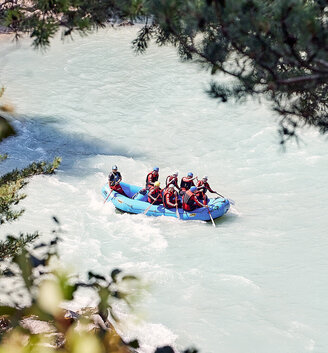 A group of people wearing helmets and life jackets is paddling a blue inflatable boat down a river framed by green branches.