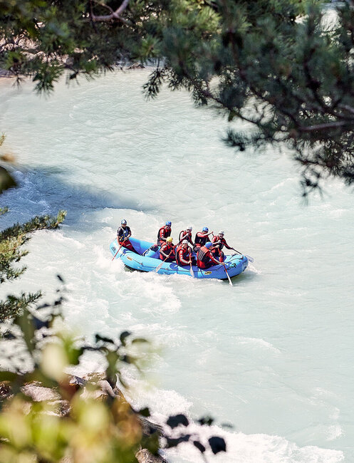 Eine Gruppe von Menschen, die Helme und Schwimmwesten tragen, paddelt in einem blauen Schlauchboot einen Fluss hinunter, der von grünen Ästen eingerahmt wird.