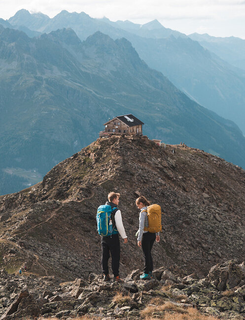 Two hikers with backpacks stand on a rocky mountain ridge, holding hands and looking at each other. A small hut sits atop the summit behind them with mist-shrouded mountains in the background.