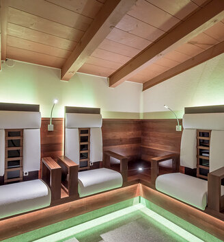 Three modern wooden infrared armchairs with white cushions are positioned in a corner beneath a wooden ceiling, illuminated by soft green accent lighting and individual reading lamps.