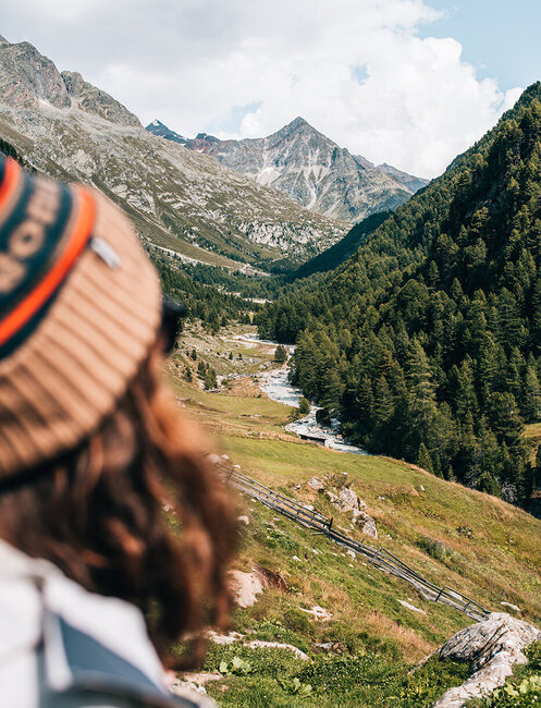 A person wearing a brown hat looks out over a picturesque mountain valley featuring a river, green hills, trees and distant peaks under a partly cloudy sky.
