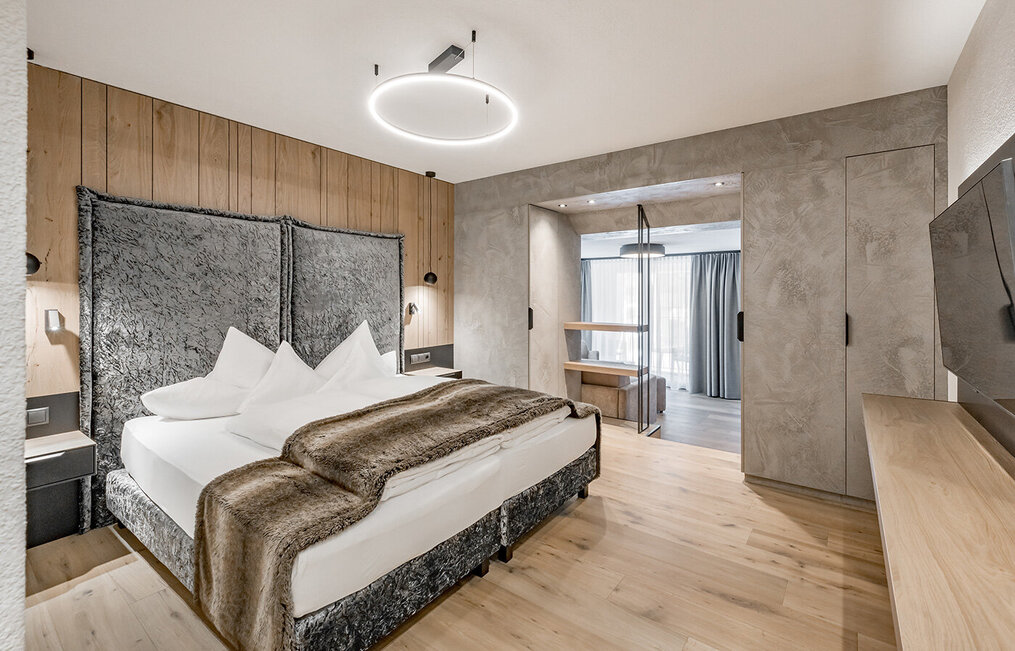 A modern hotel room featuring a large bed, a faux-fur throw, wooden flooring, a wood-accented wall, a TV, a built-in wardrobe and a seating area by large windows with sheer curtains. The ceiling light adds a contemporary touch to the room.