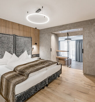 A modern hotel room featuring a large bed, a faux-fur throw, wooden flooring, a wood-accented wall, a TV, a built-in wardrobe and a seating area by large windows with sheer curtains. The ceiling light adds a contemporary touch to the room.