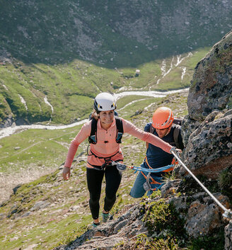 Two people wearing helmets and climbing harnesses are climbing a rocky mountain using a safety rope with a green valley and a winding river below them.