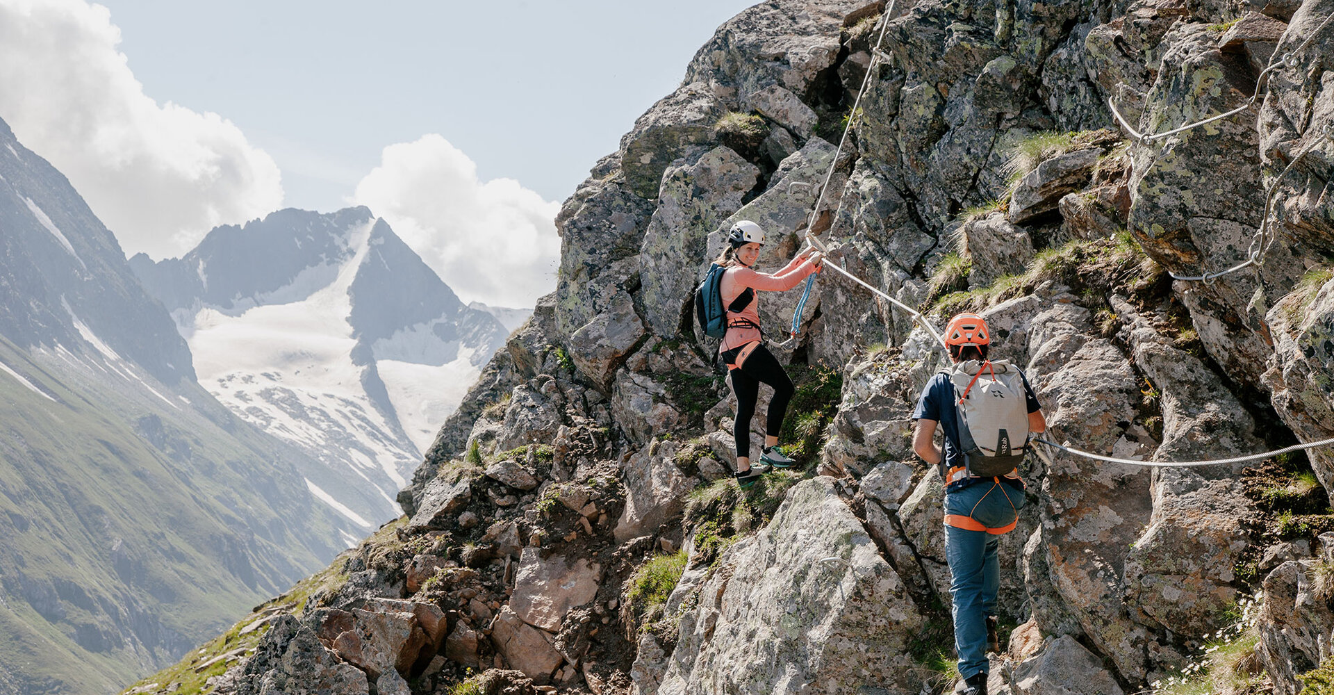 Two people wearing helmets and backpacks climb a rocky mountain path using ropes. Snow-covered peaks and a partly cloudy sky in the background.