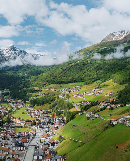 A picturesque village with many houses, green hills and dense forests, surrounded by snow-capped peaks under a partly cloudy sky.