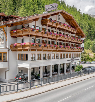 From the other side of the street, one can see a large hotel with flower-adorned wooden balconies set against a backdrop of green pines and a partly cloudy sky.
