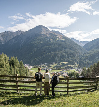 Three hikers with backpacks stand next to a wooden fence, looking out over a picturesque valley with a village surrounded by green forests and mountains under a partly cloudy sky.