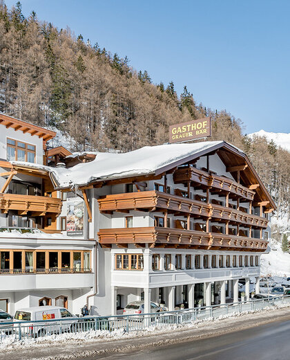 An Alpine-style hotel with balconies and snow-covered roofs, situated on a street in a snowy mountain village, with trees and mountains in the background.