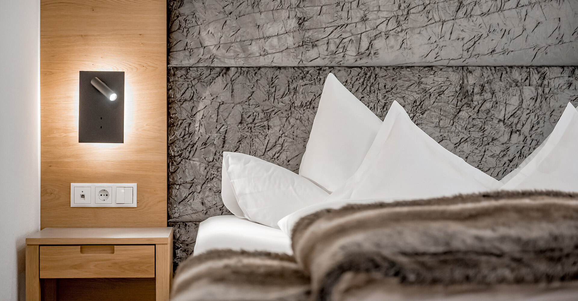 A modern hotel room features a bed with white pillows and a fur throw, a wooden nightstand, a wall-mounted reading lamp and a textured gray headboard.