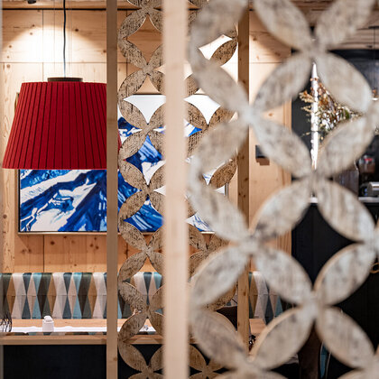 A cozy restaurant interior featuring a red pendant light, a patterned wooden partition element, seating alcoves with geometric upholstery and a blue-and-white abstract painting on the wall.