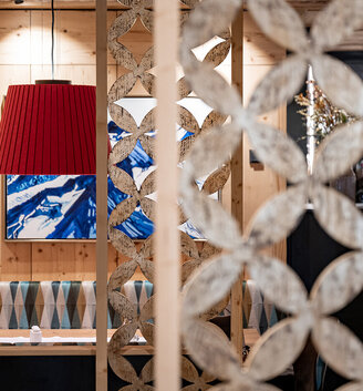 A cozy restaurant interior featuring a red pendant light, a patterned wooden partition element, seating alcoves with geometric upholstery and a blue-and-white abstract painting on the wall.