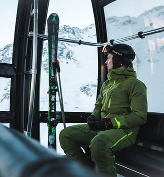 A person wearing green ski gear and a helmet is sitting in a gondola, looking out at the snow-covered mountains. The skis are standing upright on the cabin's outside.