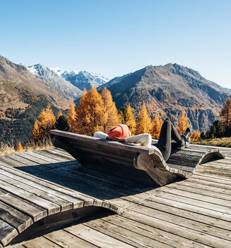 A person relaxes on a wooden deck chair on a terrace overlooking trees in autumnal colors and snow-capped mountains under a brilliant blue sky.