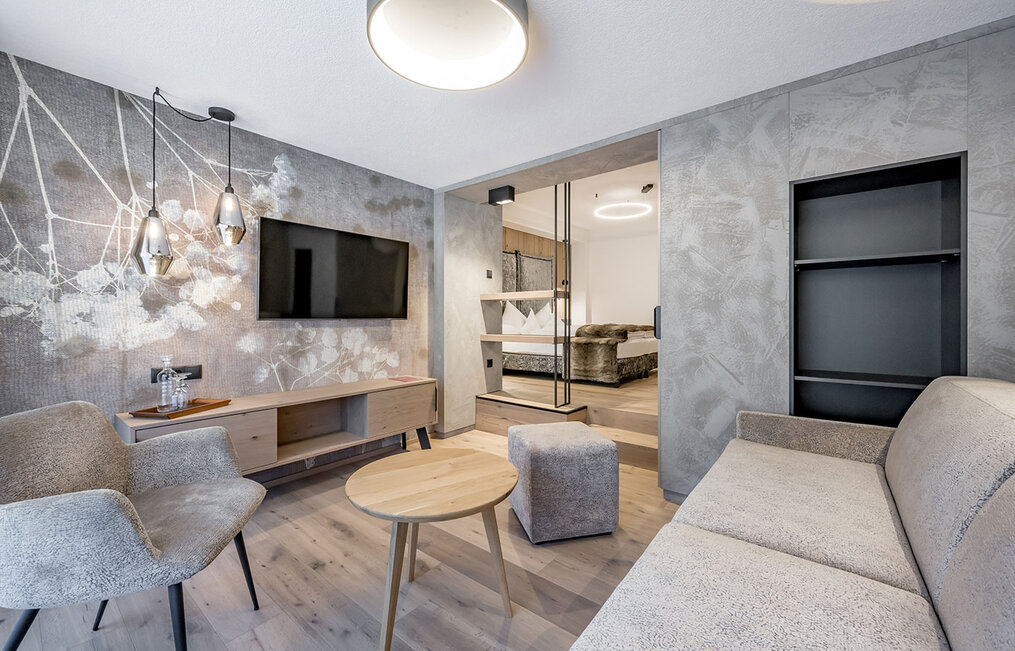 A modern living room with light gray walls, a sofa, an armchair, a round wooden coffee table, a wall-mounted TV and pendant lights. A bedroom with a bed behind an open doorway.