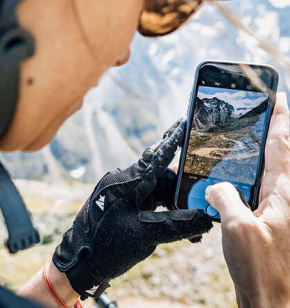 A person wearing gloves and a helmet is photographing a picturesque mountain landscape with their smartphone. On the smartphone screen there are mountains and a trail in the background.