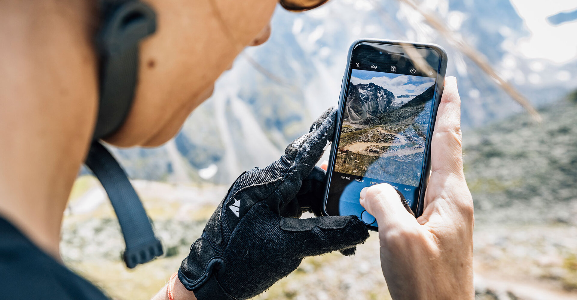 A person wearing gloves and a helmet is photographing a picturesque mountain landscape with their smartphone. On the smartphone screen there are mountains and a trail in the background.