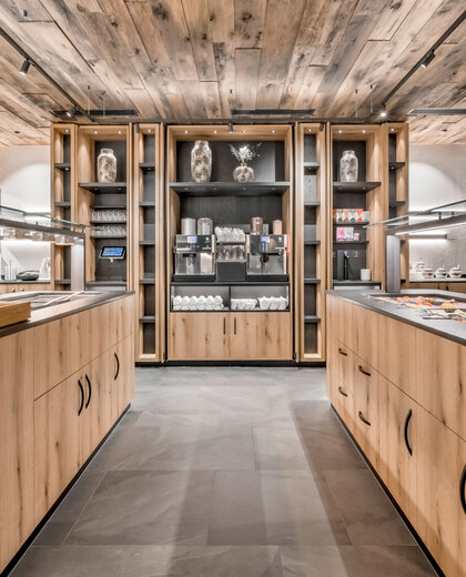 Moderner Buffetbereich mit Holzschränken, Regalen mit dekorativen Vasen, Kaffeemaschinen, Tellern, Tassen und Frühstückszubehör auf den Theken, unter warmer Beleuchtung und einer Decke aus Naturholz.