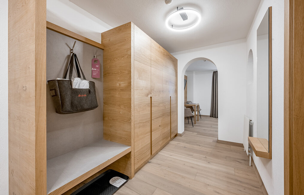 Modern hallway with wooden floors and built-in closets. An open closet area contains a grey felt bag and a clothes hanger. A hallway leads to the living area. The walls are white, the room is brightly illuminated by a round ceiling light.