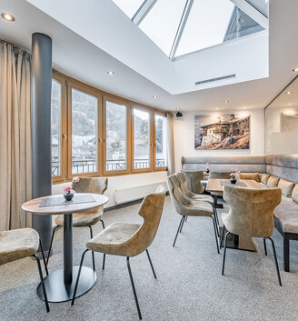 Modern lounge with grey chairs, small round tables, a long upholstered bench, large windows, a skylight and a view of the mountains. Soft lighting and a cozy, inviting atmosphere. Flowers on the tables and artwork on the walls.