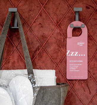 A pink door hanger and a bag containing towels hang on hooks in front of a red quilted wall.