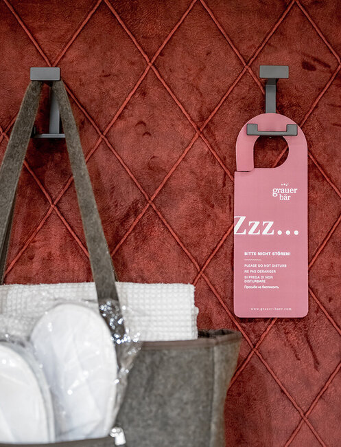 A pink door hanger and a bag containing towels hang on hooks in front of a red quilted wall.